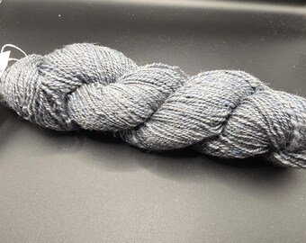 Puede incluir: Un ovillo de hilo gris con sutiles motas azules y blancas. El hilo está fuertemente torcido, creando una apariencia texturizada. Ideal para proyectos de tejido o ganchillo, este hilo ofrece una paleta de colores neutros.