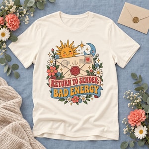 Könnte beinhalten: Ein cremefarbenes T-Shirt mit einer grafischen Illustration einer lächelnden Sonne, einer Mondsichel und einem versiegelten Briefumschlag. Der Text "RETURN TO SENDER: BAD ENERGY" steht auf einem blauen Banner, umgeben von Blumen und Sternen.