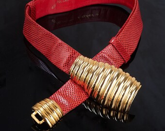 May include: A red leather belt with a gold buckle. The buckle is made of multiple rings that are stacked on top of each other. The belt is signed "Judith Leiber's" on the inside.