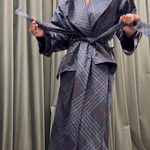 May include: A two-piece outfit featuring a jacket and a skirt. The jacket has a notched collar and a matching belt. The skirt is a long, flowing style. The fabric has a plaid pattern in shades of blue, gray, and brown.