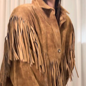 May include: A tan suede jacket featuring a pointed collar, a button-down front with visible buttons, and long fringe detailing across the chest, shoulders, and sleeves. The garment is constructed from a soft, textured material.