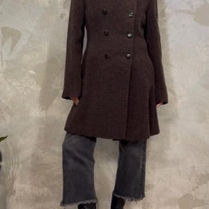 May include: A brown double-breasted wool coat with a black velvet collar. The coat has a fitted silhouette and a flared hem. The model is wearing the coat with grey jeans and black heeled boots.