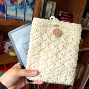 Puede incluir: Funda de lector electrónico de ganchillo color crema con cierre de botón de madera. La funda tiene un patrón texturizado y un lazo en la parte superior. El lector electrónico muestra texto de un libro. El botón de madera dice "handmade with love".