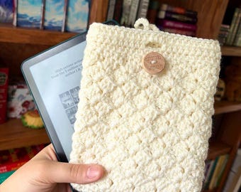 May include: Cream-colored, crocheted e-reader case with a wooden button closure. The case has a textured pattern and a loop at the top. The e-reader displays text from a book. The wooden button says "handmade with love".
