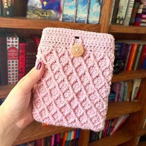 May include: A light pink crochet pouch featuring a textured diamond pattern across the front. The rectangular sleeve is secured at the top centre with a single round wooden button that has the word handmade engraved on its surface.