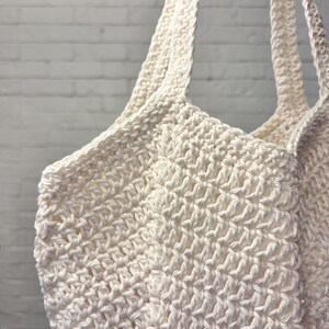May include: Off-white crochet tote bag with long straps. The bag is made of a thick, textured yarn, with a visible stitch pattern. The bag's handles are also crocheted, and the background is a white brick wall.