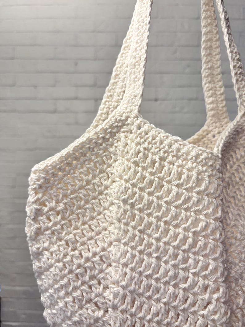 May include: Off-white crochet tote bag with long straps. The bag is made of a thick, textured yarn, with a visible stitch pattern. The bag's handles are also crocheted, and the background is a white brick wall.