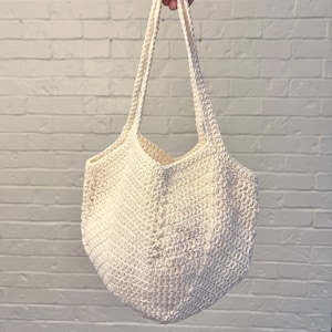 May include: Off-white crochet tote bag with long straps. The bag has a geometric shape and a textured, open weave pattern. The bag is held up against a white brick wall.