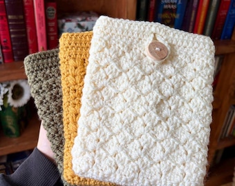 May include: Three handmade crochet book covers in cream, mustard yellow, and olive green. The cream cover has a wooden button closure. The covers are made with a textured stitch pattern and are displayed in front of a bookshelf.