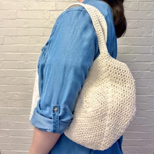 May include: Cream-colored crochet bag with a rounded shape and a shoulder strap. The bag has a textured, open weave design. The strap is made of the same material as the bag. The bag is being carried over the shoulder.