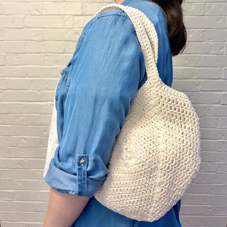 May include: Cream-colored crochet bag with a rounded shape and a shoulder strap. The bag has a textured, open weave design. The strap is made of the same material as the bag. The bag is being carried over the shoulder.