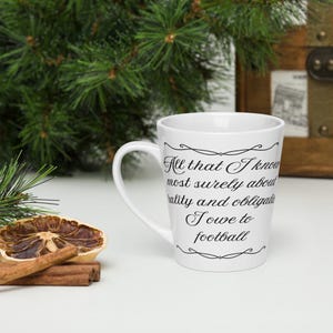 May include: A white ceramic latte mug featuring black cursive text that reads, All that I know most surely about morality and obligations I owe to football, framed by decorative black flourishes at the top and bottom of the quote.