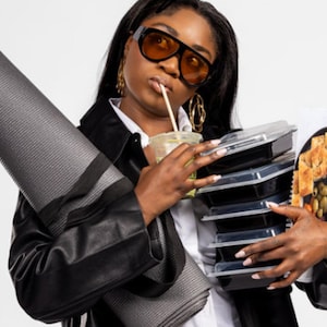 May include: A woman wearing sunglasses and a black leather jacket holds a rolled-up yoga mat, a drink, and a stack of black food containers. The image includes the text "Soulful Snacking" and "actually written by a human."