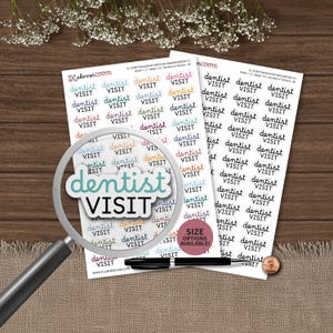 May include: Two sheets of planner stickers featuring the text "dentist VISIT" in various colors and black. A magnifying glass is positioned over one sticker, and a black pen and a copper penny are placed on the white sticker sheets.