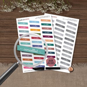 May include: Two sheets of rectangular planner stickers featuring the word cancelled in white script. One sheet contains multi-colored stickers, while the other features grey stickers. A magnifying glass, a black pen, and a copper penny are present.