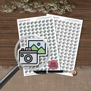 Könnte beinhalten: Zwei Bögen mit Planer-Aufklebern, die kleine Kamera- und Landschaftssymbole zeigen. Ein Bogen enthält farbige Symbole, der andere schwarz-weiße. Eine Lupe fokussiert ein einzelnes Kamerasymbol. Text: plannerzoom und size options available.
