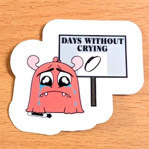 May include: A die-cut sticker featuring a pink cartoon monster with large teary eyes and blue tears streaming down its face. The monster holds a black marker next to a light blue sign that reads DAYS WITHOUT CRYING with a large handwritten zero.