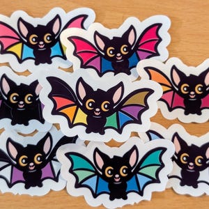 May include: A collection of cartoon bat stickers. Each bat has a black body, pink ears, and colorful wings. The wings feature a rainbow of colors, including red, orange, yellow, green, blue, and purple. The bats have a smiling face.