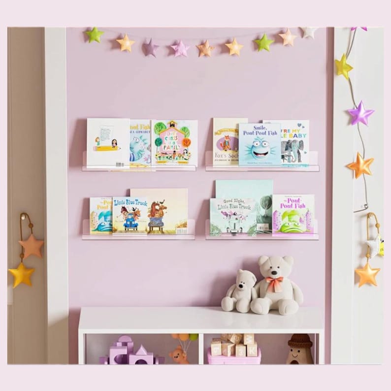 May include: Four horizontal white floating shelves mounted on a light purple wall, displaying various children's books. A string of colorful fabric stars hangs above the shelves. A white console table below holds two plush teddy bears and toys.