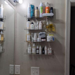 May include: Four clear acrylic wall-mounted shelves holding various skincare and hair care products, including bottles, tubes, jars, and a small stack of folded gray cloths. The items are arranged in rows on the shelves against a plain wall.