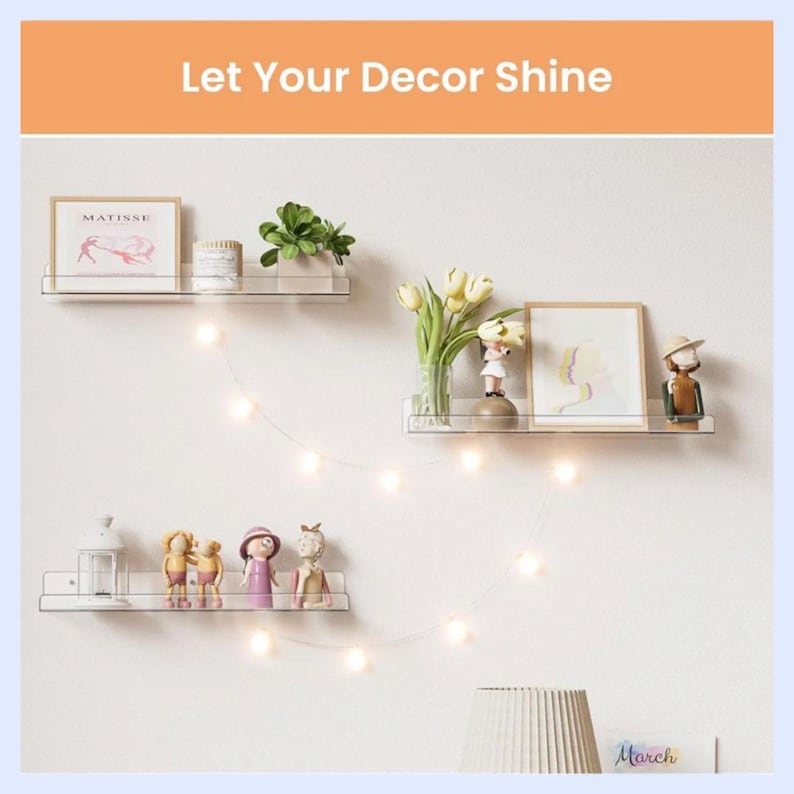 May include: Three clear acrylic floating wall shelves arranged in a staggered pattern. The shelves hold framed art, small potted plants, decorative figurines, and a white lantern. A string of warm white LED fairy lights hangs between the shelves.