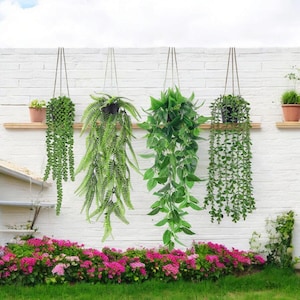 May include: Four hanging planters with green trailing foliage suspended from a horizontal wooden shelf against a white brick wall. The plants include a string of pearls, a fern, a broad-leafed vine, and a succulent vine, all in dark pots.