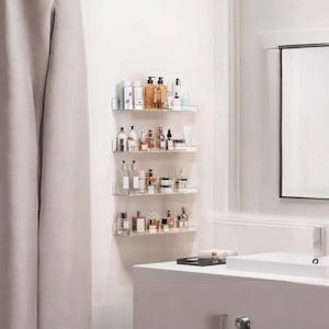 May include: A set of four clear acrylic wall-mounted floating shelves arranged vertically. Each shelf holds various glass bottles, jars, and containers of skincare and cosmetic products, providing organized storage for bathroom essentials.