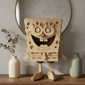 May include: A light-colored wooden cutout figure of a cartoon character with a rectangular body, large circular eyes, a wide smiling mouth with two teeth, thin arms, and two legs with flat feet, standing upright with a layered, textured appearance.