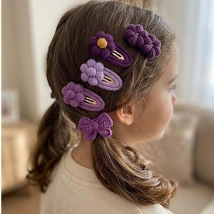 May include: A collection of purple crochet hair clips, including flower and bow designs, are displayed in a young girl's hair. The clips have gold-colored metal clasps. One clip features a yellow center.