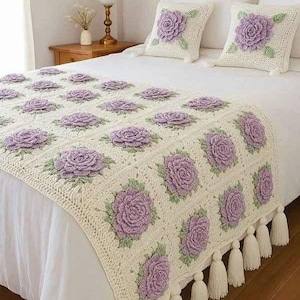 Pode incluir: Um caminho de cama de crochê na cor creme e duas almofadas quadradas correspondentes, cada uma com um motivo de rosa lavanda tridimensional com folhas verdes. O caminho é finalizado com longas borlas na cor creme na borda inferior.