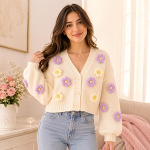 May include: Cream-coloured cardigan sweater decorated with crocheted daisy appliqués in purple and yellow. The V-neck sweater has long sleeves and a button-down front. The sweater is paired with light blue jeans.