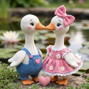 May include: Two crocheted goose dolls, one in blue overalls with a pink heart, the other in a pink dress with a bow. They are kissing, with a small pink heart between them. The dolls are on a stone surface with a pond in the background.
