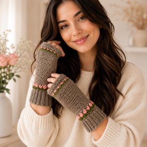 May include: A pair of brown fingerless gloves with a ribbed cuff and a decorative band of pink flowers and green leaves. The gloves are made of a knitted material and are shown on a person wearing a cream-colored sweater.