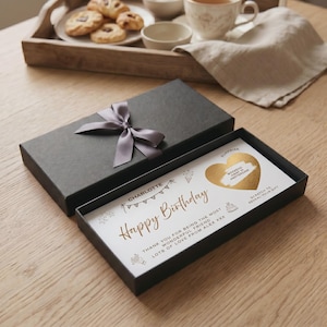 Può includere: Una scatola regalo rettangolare nera con un fiocco di nastro di raso grigio, contenente un biglietto bianco con testo dorato che recita Happy Birthday, Charlotte, e un cuore dorato da grattare con il testo Weekend Away in Amsterdam.