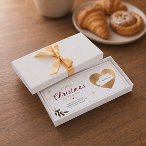 Può includere: Una scatola regalo rettangolare bianca con un nastro dorato, contenente un biglietto con la scritta A LITTLE Christmas GIFT FOR YOU, un cuore dorato da grattare con il testo ST IVES GETAWAY, illustrazioni di agrifoglio e il testo WITH LOVE EMMA X.