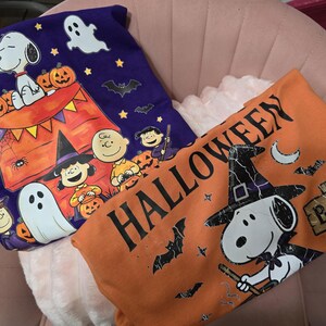 May include: A purple t-shirt featuring Snoopy, Charlie Brown, and Lucy with pumpkins, ghosts, and bats, alongside an orange t-shirt with the text HALLOWEEN, a graphic of Snoopy wearing a witch hat, bats, and a wooden sign that reads BOO.