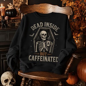 Könnte beinhalten: Ein schwarzes Sweatshirt mit Rundhalsausschnitt und einer Grafik eines Skeletts, das in einem Sarg sitzt und eine dampfende Tasse mit Totenkopf hält. Der Text lautet DEAD INSIDE BUT CAFFEINATED, ergänzt durch Fledermaus- und Sternmotive.