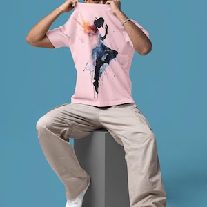 May include: A light pink short-sleeved t-shirt featuring a graphic print of a dancing figure in a hat. The silhouette is rendered in black with vibrant watercolor splashes of orange, blue, and purple emanating from the figure's upper body.