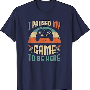 Op de afbeelding: Een marineblauw t-shirt met korte mouwen en een grafische print met een retro zonsondergang, het silhouet van een zwarte gamecontroller en de tekst "I PAUSED MY GAME TO BE HERE" in een meerkleurig, verweerd lettertype met kleine sterren.