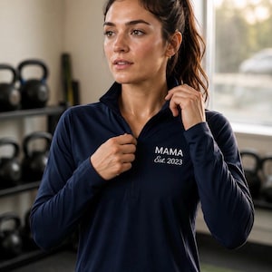 Puede incluir: Top deportivo azul marino de manga larga con cuello con cremallera. El texto "MAMA Est. 2023" está impreso en blanco en el pecho. La parte superior está hecha de un material ligero y transpirable, adecuado para entrenamientos.