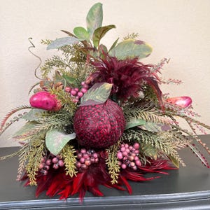 May include: A decorative floral arrangement featuring a central, textured, deep red sphere surrounded by greenery, berries, and burgundy feathers. The arrangement includes various shades of green leaves and small, round, pink berries, creating a rich, autumnal aesthetic.