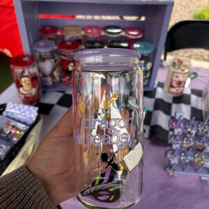 May include: A clear glass can-shaped tumbler with a purple lid. The tumbler features the text "dragon reader" with illustrations of a dragon, book, and lightning bolt. Other decorated glass jars are visible in the background.