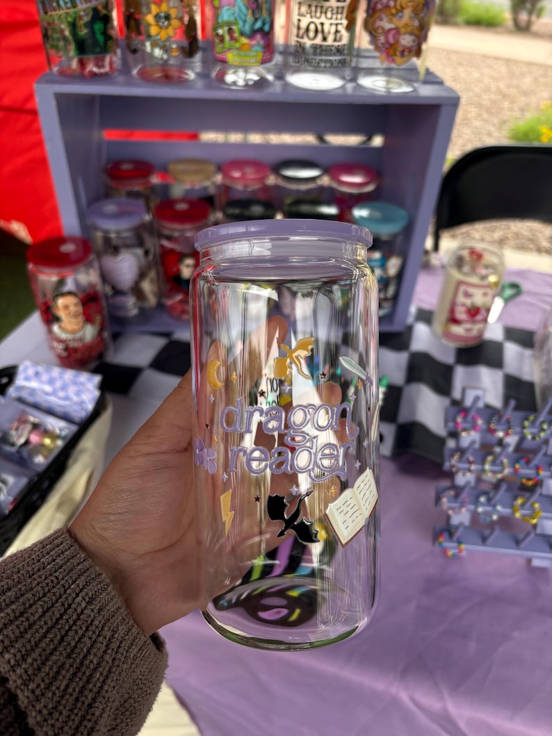 May include: A clear glass can-shaped tumbler with a purple lid. The tumbler features the text "dragon reader" with illustrations of a dragon, book, and lightning bolt. Other decorated glass jars are visible in the background.