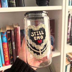 May include: A clear glass tumbler with a black lid. The tumbler features a black and cream-colored graphic with the words "STILL EMO" and a red broken heart. The tumbler is held in front of a bookshelf.