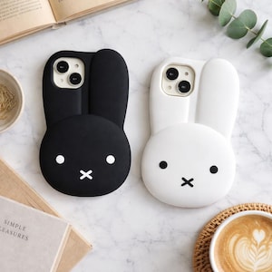 Op de afbeelding: Twee telefoonhoesjes in de vorm van een konijn, één zwart en één wit. Elke hoes heeft een rond gezicht met een kruis als mond en stipogen. De zwarte hoes heeft witte ogen, terwijl de witte hoes zwarte ogen heeft.