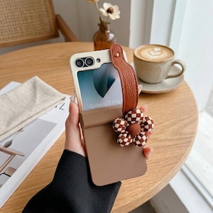 May include: A brown phone case with a leather strap and a checkered flower accent. The phone case is designed for a Galaxy Z Flip phone. The phone screen displays the words "Galaxy Z Flip".