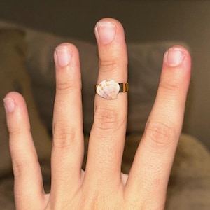 May include: A gold-toned metal ring featuring a small, natural seashell attached to the center of the band. The seashell has a smooth, fan-shaped appearance with light tan and white coloration, contrasting against the polished gold-colored metal band.