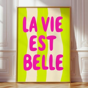 Op de afbeelding: Een ingelijste kunstprint met de tekst "LA VIE EST BELLE" in felroze, tegen een golvende achtergrond van limoengroen en crème. Het kunstwerk staat op een houten vloer, tegen een witte muur.