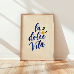 May include: A rectangular wall art print in a light wood frame featuring the blue brush-lettered text La Dolce Vita on a cream-colored background. A small yellow lemon with green leaves and a white blossom is positioned next to the text.