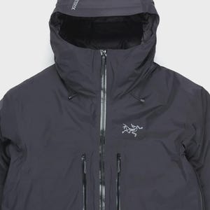 Könnte beinhalten: Eine anthrazitfarbene Kapuzenjacke mit durchgehendem Frontreißverschluss, zwei vertikalen Brusttaschen mit versiegelten Reißverschlüssen und einer verstellbaren Kapuze. Der Markenname Arc'teryx steht auf der Kapuze, das Logo auf der Brust.
